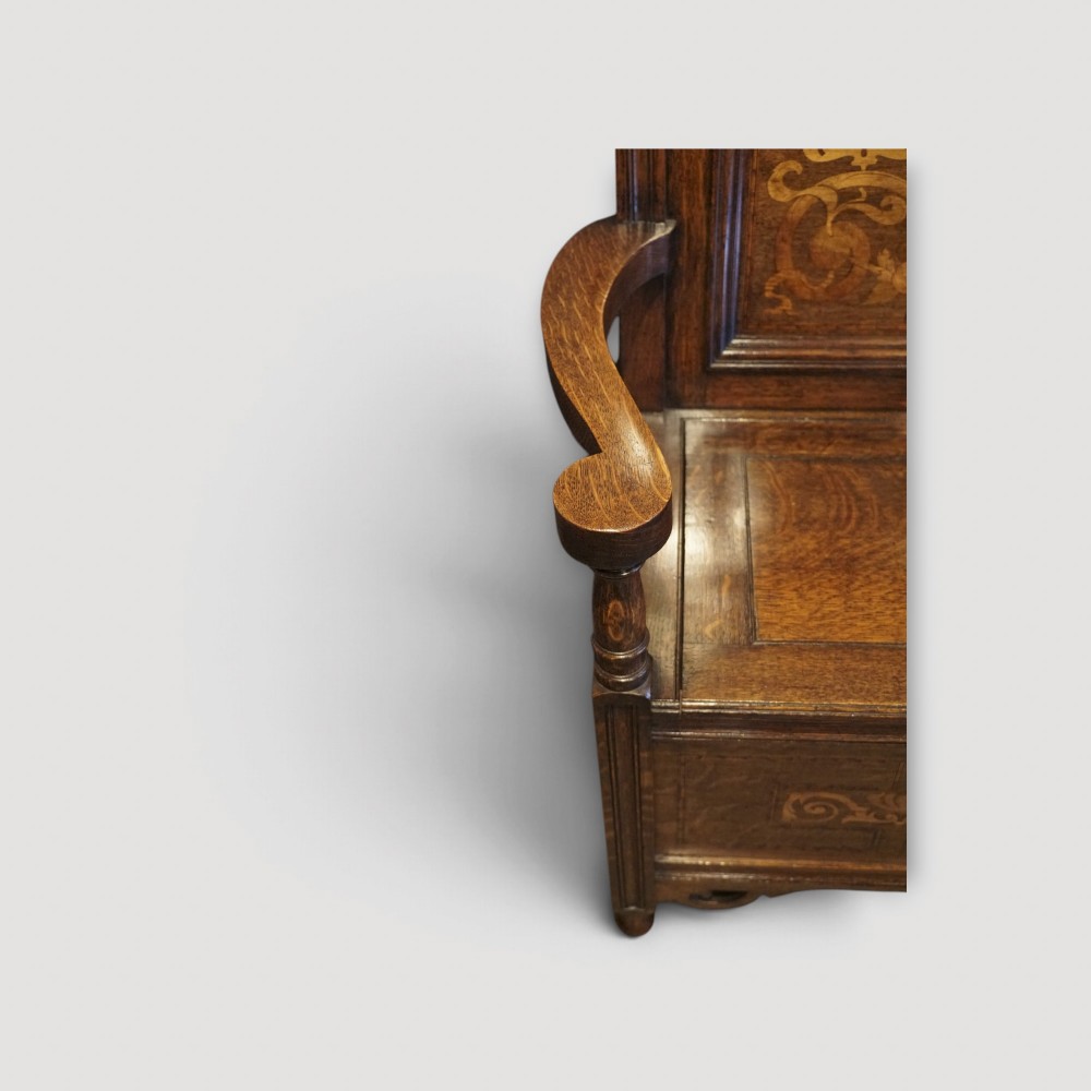 An Art Nouveau solid oak settle featuring decorative marquetry inlay across the backrest and lower front panels, curved armrests, and a lift-up storage seat. View III