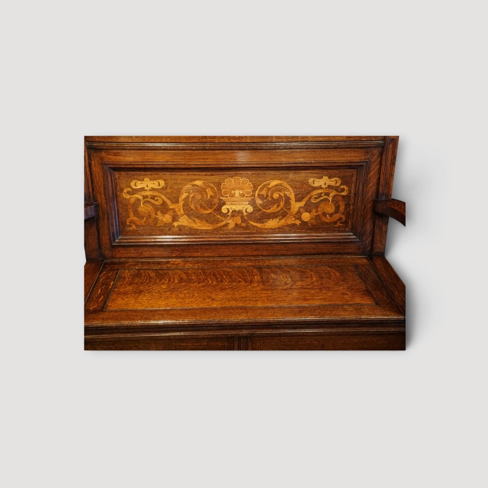 An Art Nouveau solid oak settle featuring decorative marquetry inlay across the backrest and lower front panels, curved armrests, and a lift-up storage seat. View VI