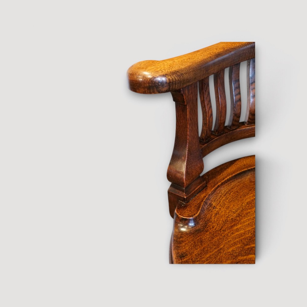 An Edwardian dark oak revolving desk chair featuring a curved slatted backrest, shaped wooden saddle seat, and four legs with castors. View II
