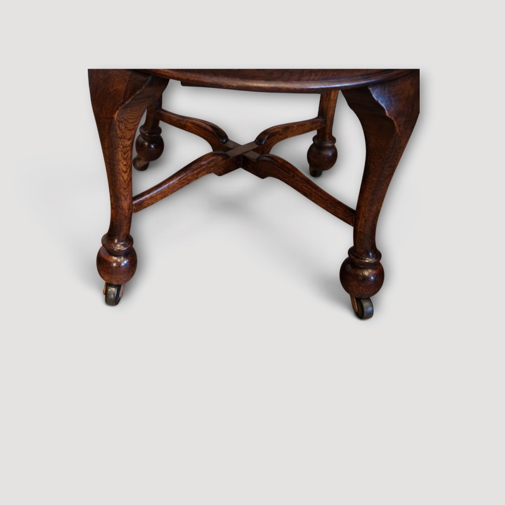 An Edwardian dark oak revolving desk chair featuring a curved slatted backrest, shaped wooden saddle seat, and four legs with castors. View V