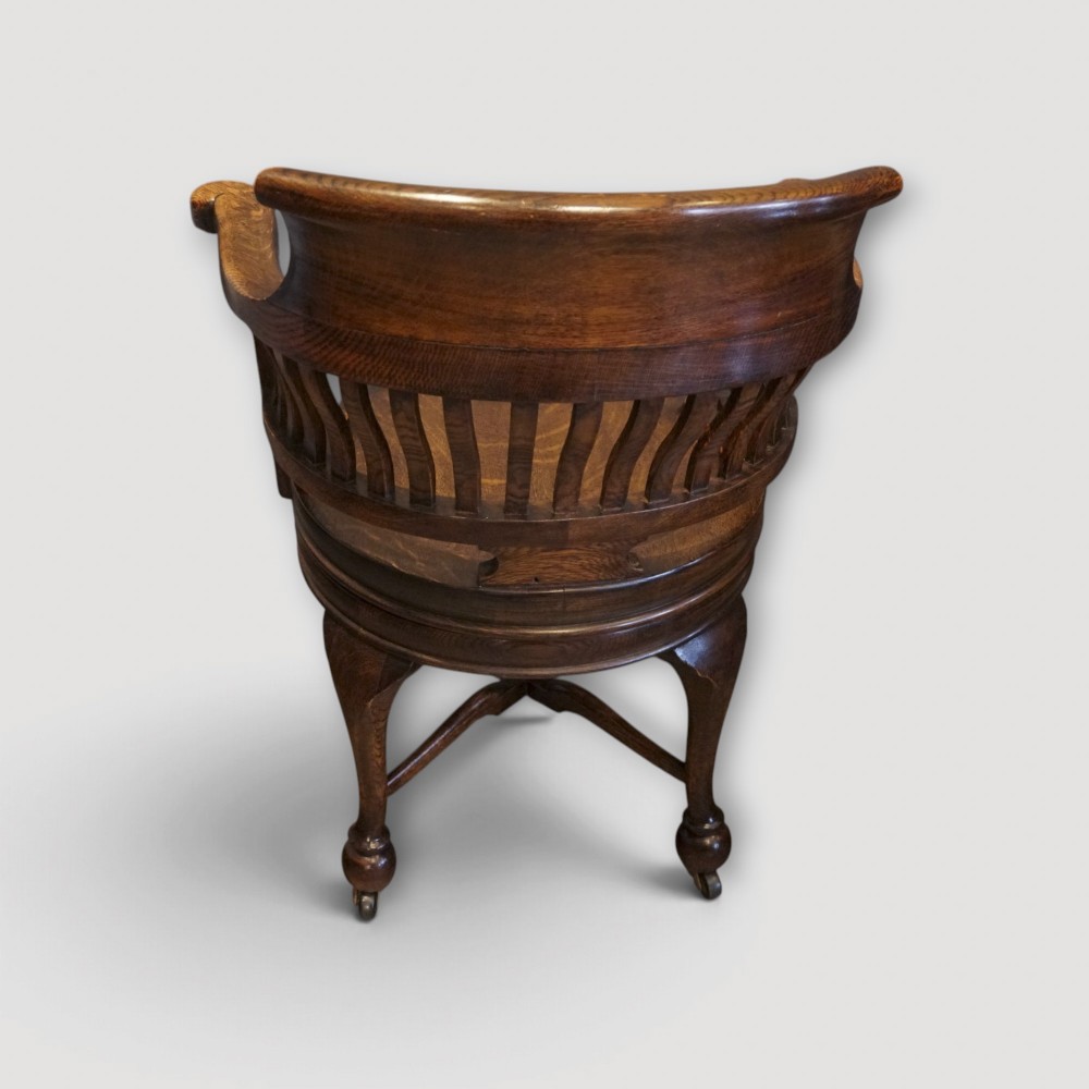 An Edwardian dark oak revolving desk chair featuring a curved slatted backrest, shaped wooden saddle seat, and four legs with castors. View VII