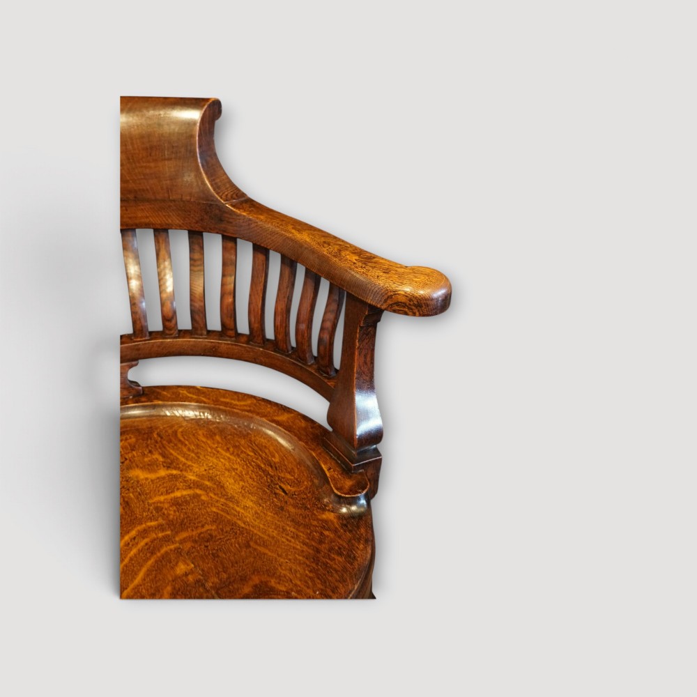 An Edwardian dark oak revolving desk chair featuring a curved slatted backrest, shaped wooden saddle seat, and four legs with castors. View IX