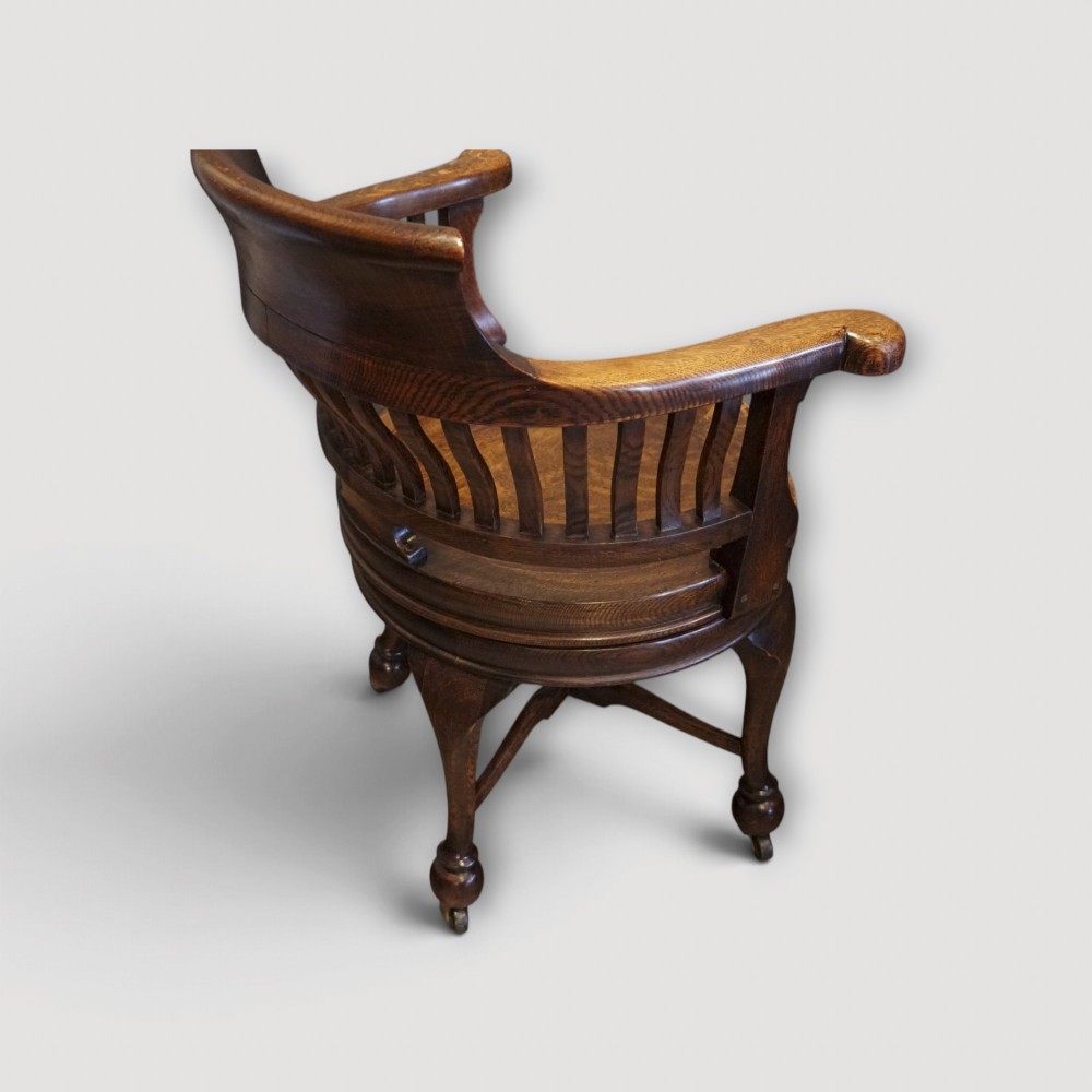 An Edwardian dark oak revolving desk chair featuring a curved slatted backrest, shaped wooden saddle seat, and four legs with castors. View X