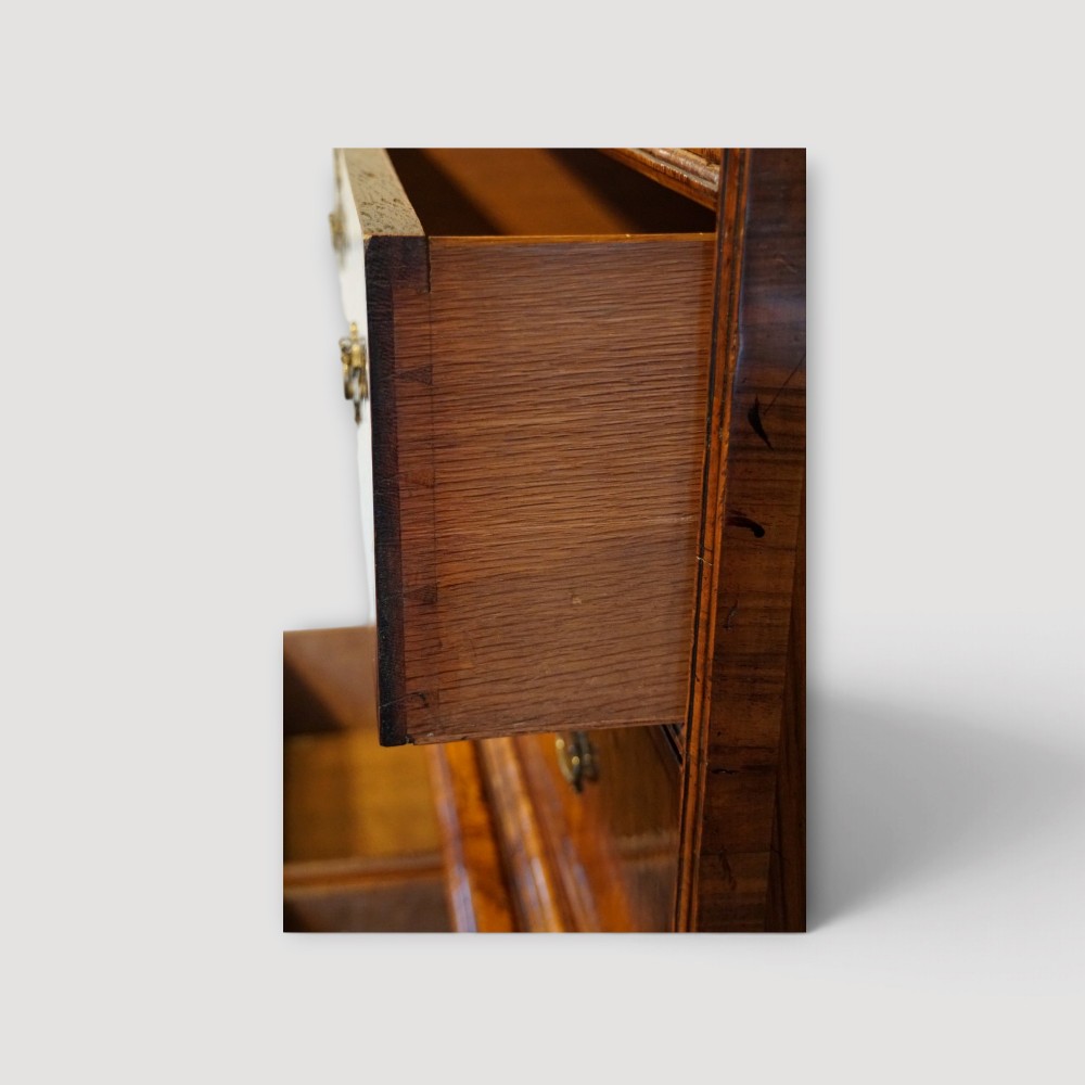 An antique Queen Anne walnut chest-on-chest in two sections featuring a multi-drawer configuration, figured grain patterns, brass drop handles, and bun feet. View V