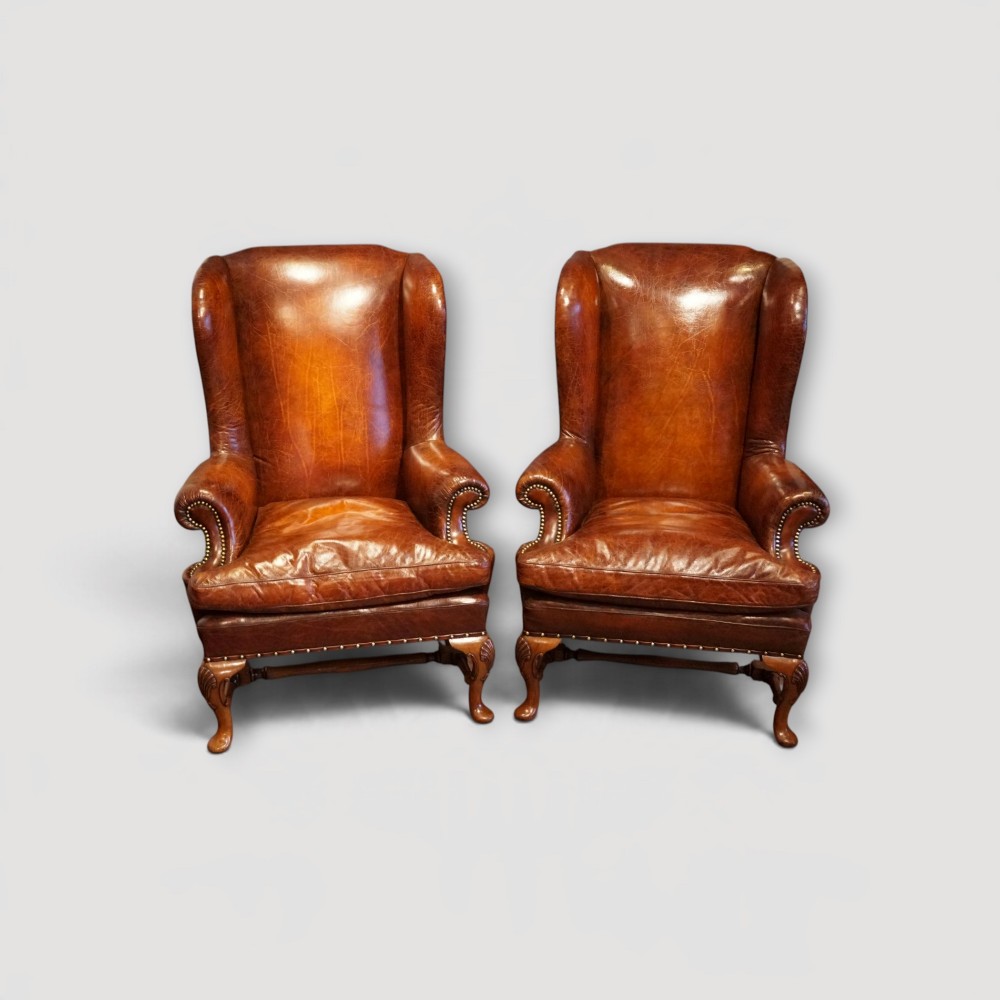 A pair of brown leather Queen Anne style wingback armchairs featuring deep seat cushions, brass stud detailing, and carved walnut cabriole legs.