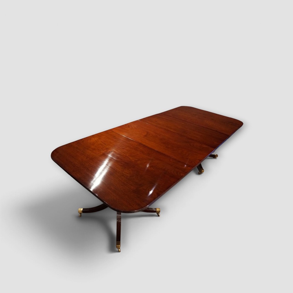An extended mahogany twin pedestal dining table featuring a polished surface, rounded corners, reeded tripod legs, and brass castors. View II
