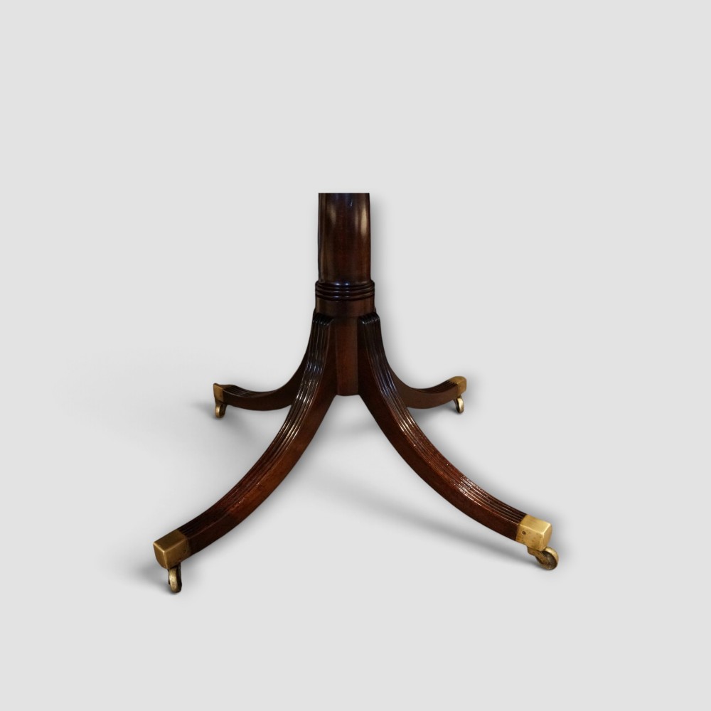 An extended mahogany twin pedestal dining table featuring a polished surface, rounded corners, reeded tripod legs, and brass castors. View III