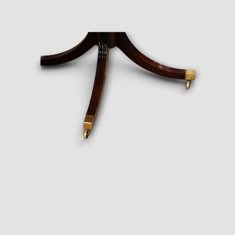 An extended mahogany twin pedestal dining table featuring a polished surface, rounded corners, reeded tripod legs, and brass castors. View IV
