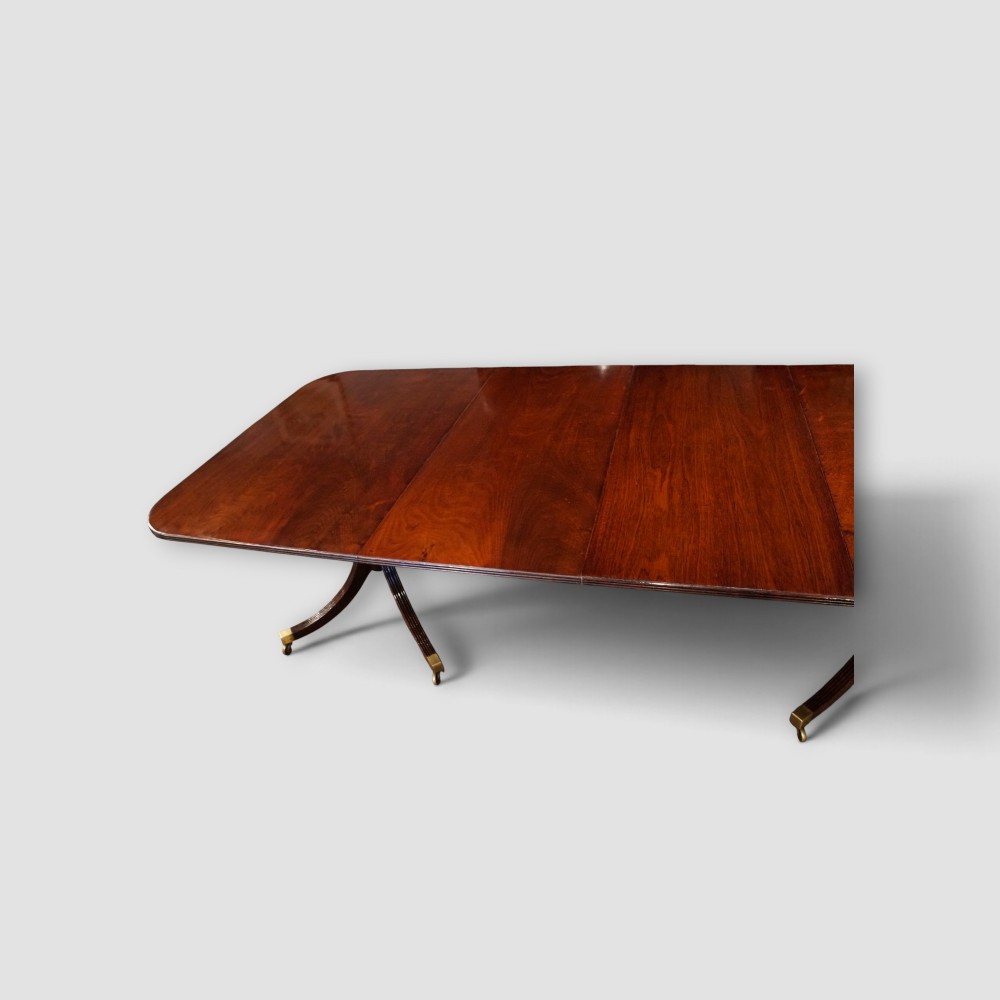 An extended mahogany twin pedestal dining table featuring a polished surface, rounded corners, reeded tripod legs, and brass castors. View IX