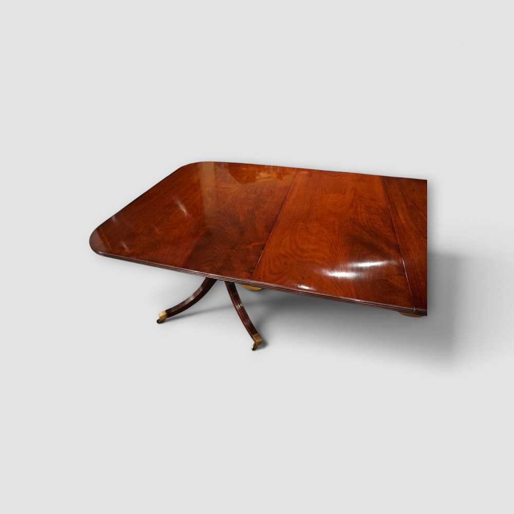 An extended mahogany twin pedestal dining table featuring a polished surface, rounded corners, reeded tripod legs, and brass castors. View X