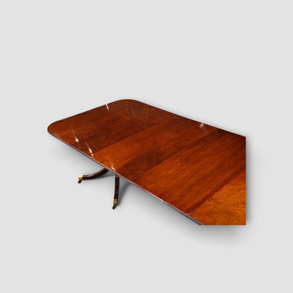 An extended mahogany twin pedestal dining table featuring a polished surface, rounded corners, reeded tripod legs, and brass castors. View XI