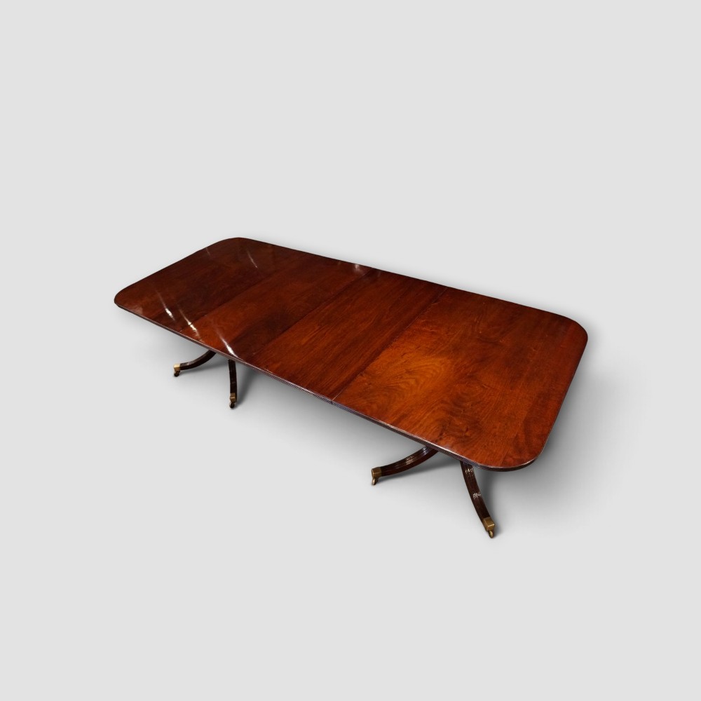 An extended mahogany twin pedestal dining table featuring a polished surface, rounded corners, reeded tripod legs, and brass castors. View XII
