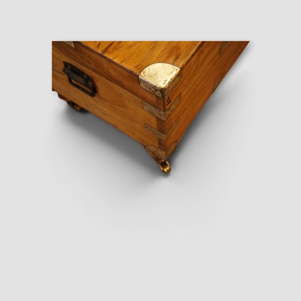 A low rectangular warm amber camphor wood campaign trunk featuring brass strap corners, flush brass handles, dual keyholes, and turned legs with castors. View II