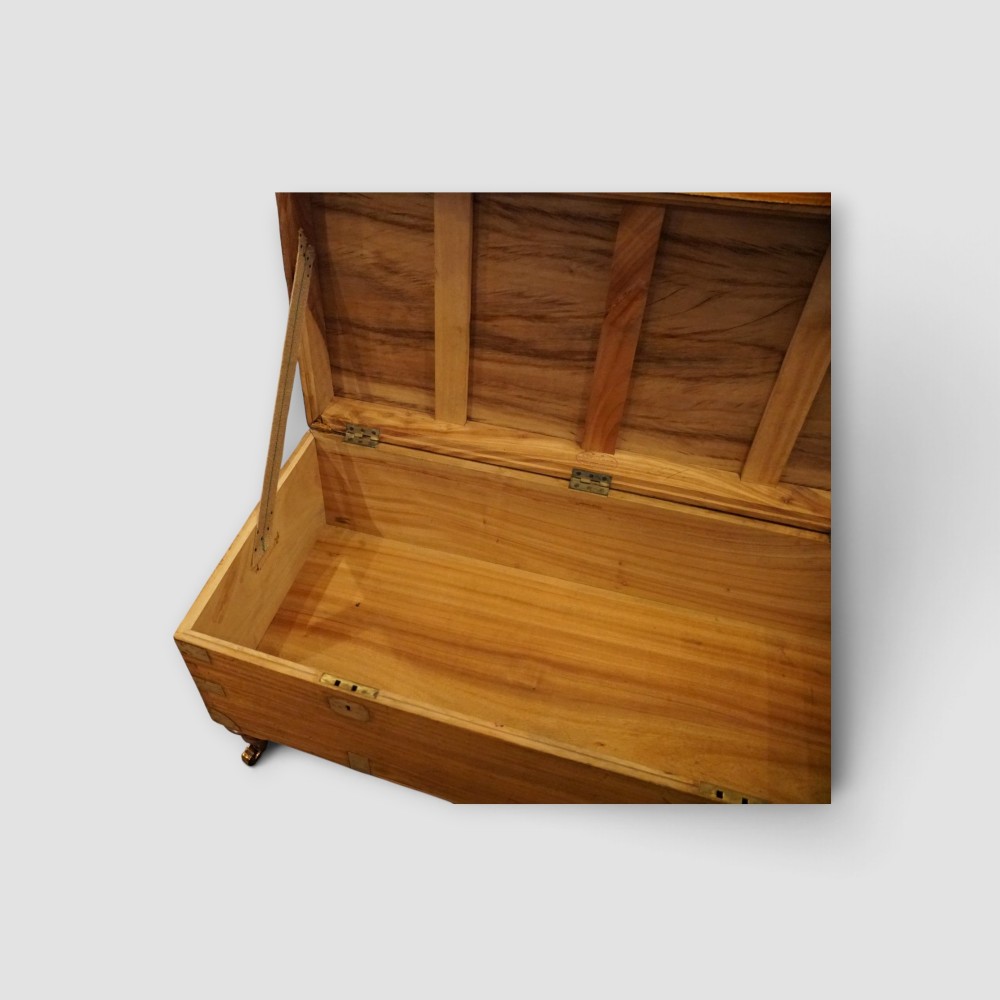 A low rectangular warm amber camphor wood campaign trunk featuring brass strap corners, flush brass handles, dual keyholes, and turned legs with castors. View IX