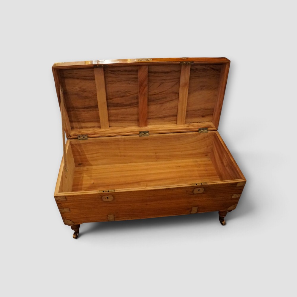 A low rectangular warm amber camphor wood campaign trunk featuring brass strap corners, flush brass handles, dual keyholes, and turned legs with castors. View XI