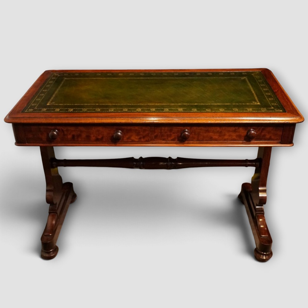 An early Victorian mahogany writing table featuring a green leather inset top with gilt tooling, two frieze drawers, a turned stretcher, and end supports.