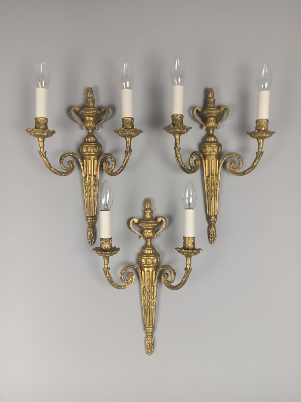 Set of three French gilded brass wall sconces featuring twin scrolling arms and urn-shaped tapered backplates.