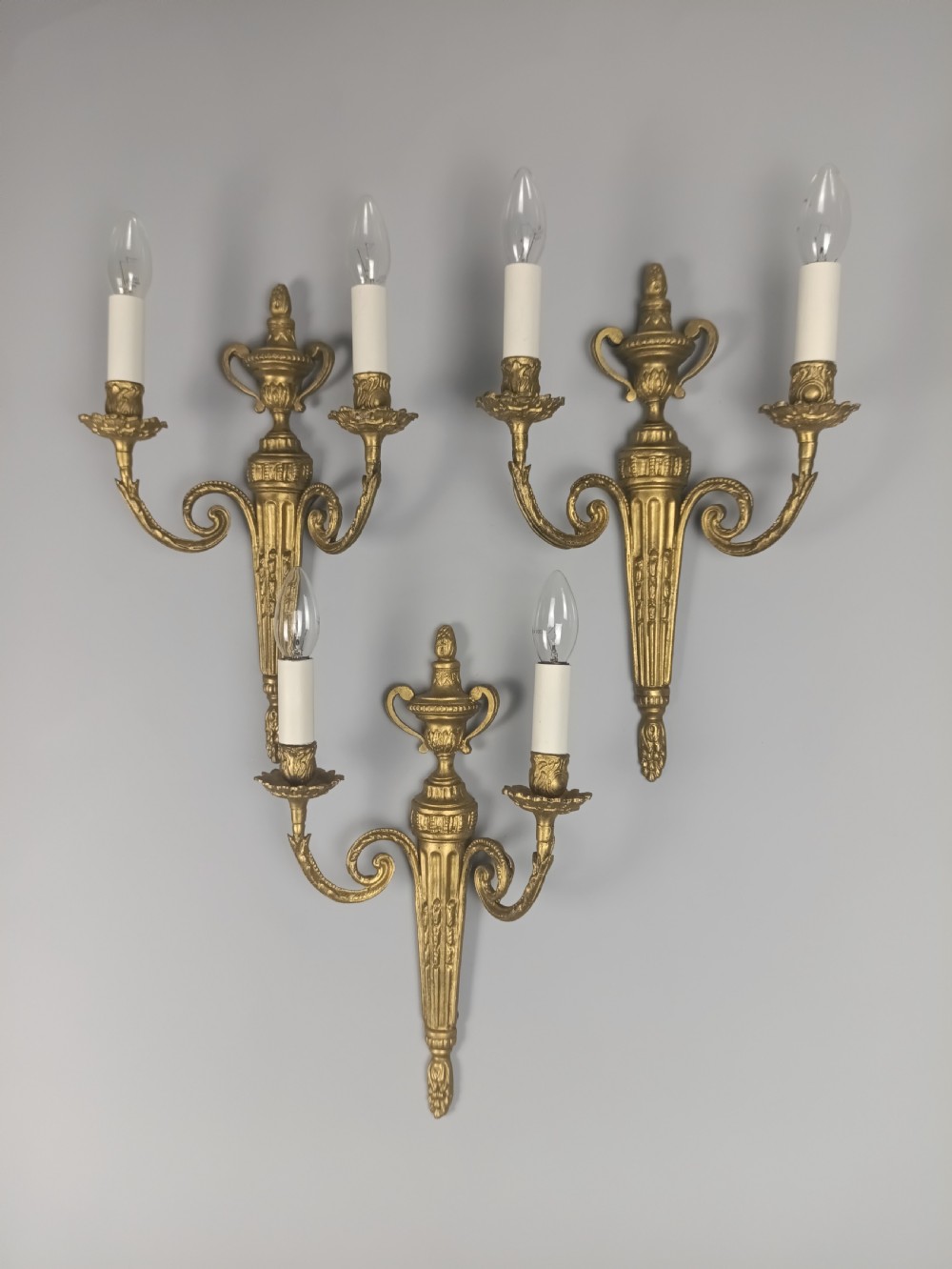 Set of three French gilded brass wall sconces featuring twin scrolling arms and urn-shaped tapered backplates. View II