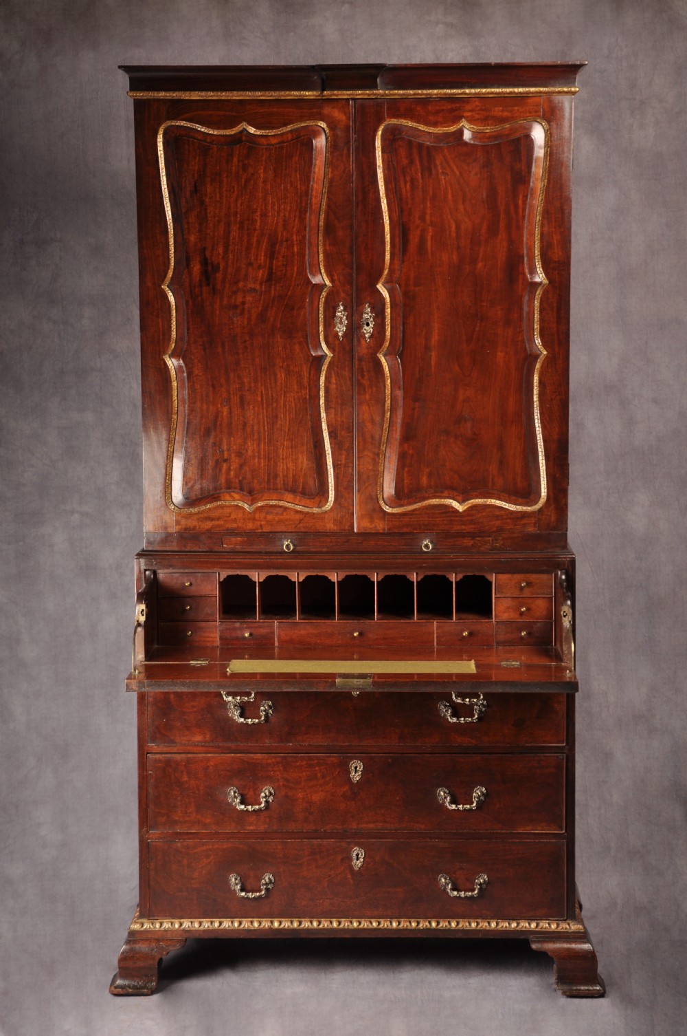 An 18th-century mahogany secretaire cabinet featuring twin upper doors with shaped gilded mouldings above an open fall-front desk with pigeonholes and three drawers on bracket feet.