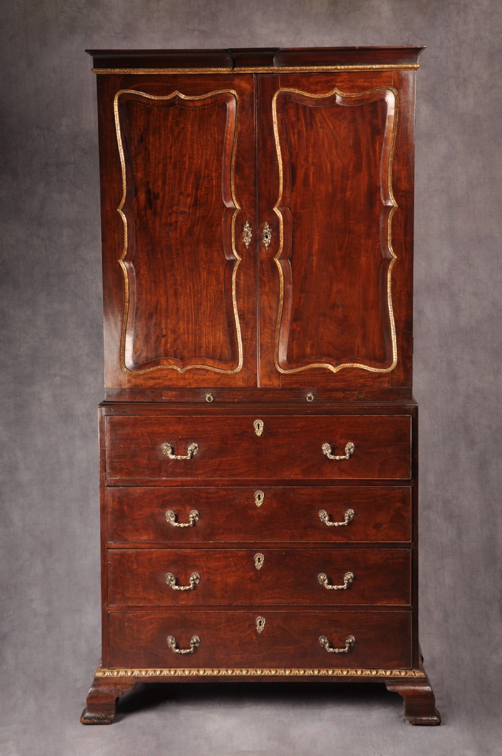 An 18th-century mahogany secretaire cabinet featuring twin upper doors with shaped gilded mouldings above an open fall-front desk with pigeonholes and three drawers on bracket feet. View II