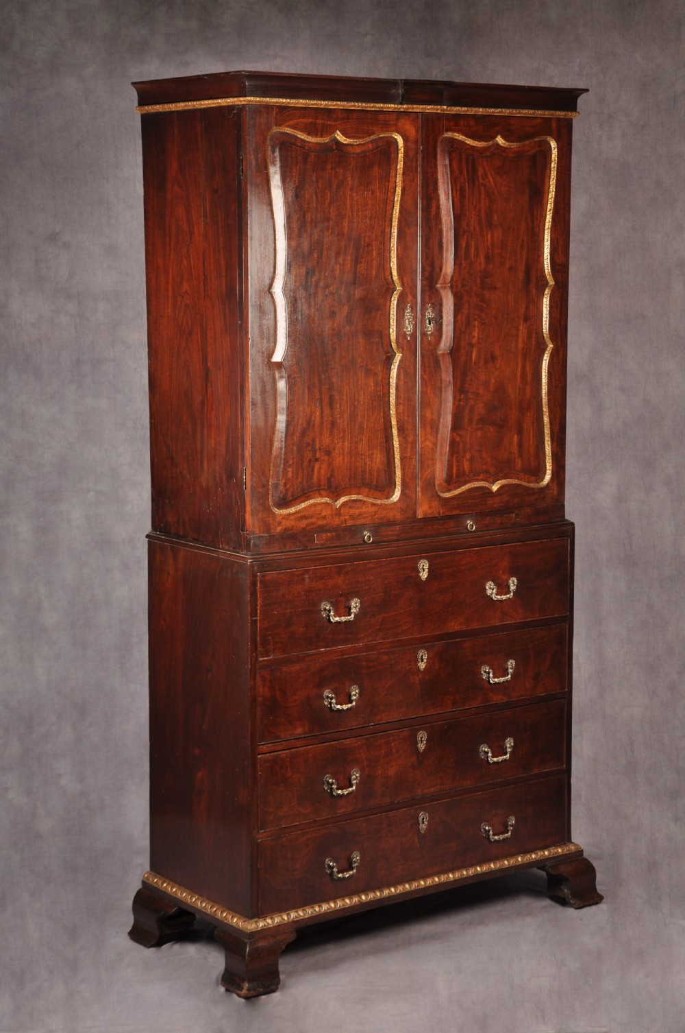 An 18th-century mahogany secretaire cabinet featuring twin upper doors with shaped gilded mouldings above an open fall-front desk with pigeonholes and three drawers on bracket feet. View XVII