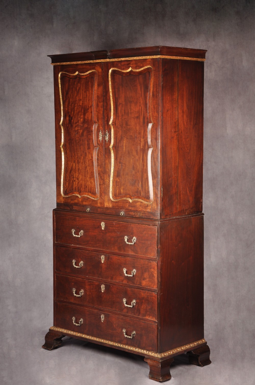 An 18th-century mahogany secretaire cabinet featuring twin upper doors with shaped gilded mouldings above an open fall-front desk with pigeonholes and three drawers on bracket feet. View XVIII