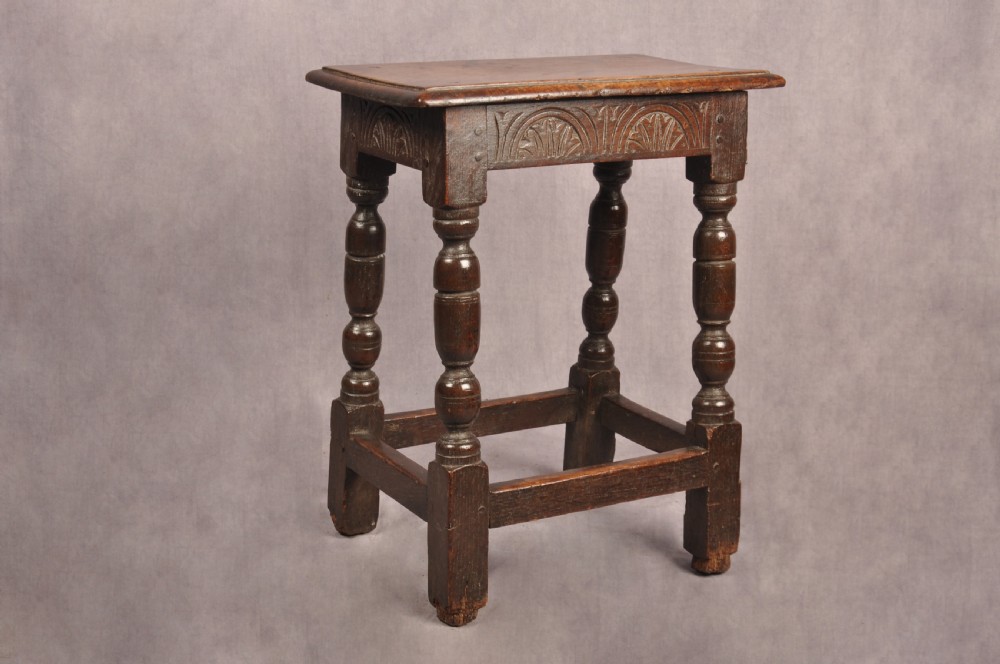 A dark oak joint stool featuring a rectangular moulded top above a carved frieze, supported by turned legs joined by low stretchers.