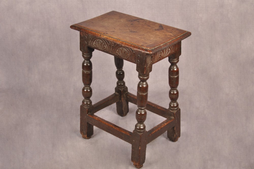 A dark oak joint stool featuring a rectangular moulded top above a carved frieze, supported by turned legs joined by low stretchers. View II
