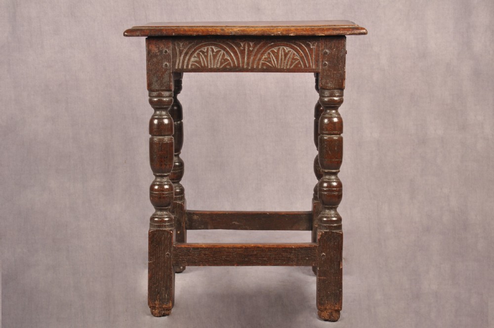 A dark oak joint stool featuring a rectangular moulded top above a carved frieze, supported by turned legs joined by low stretchers. View V