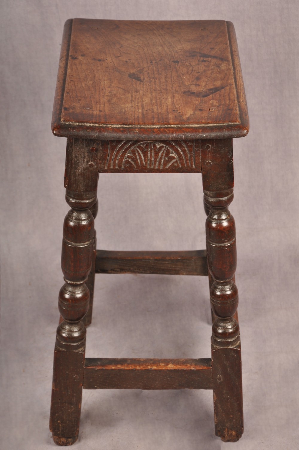 A dark oak joint stool featuring a rectangular moulded top above a carved frieze, supported by turned legs joined by low stretchers. View VI