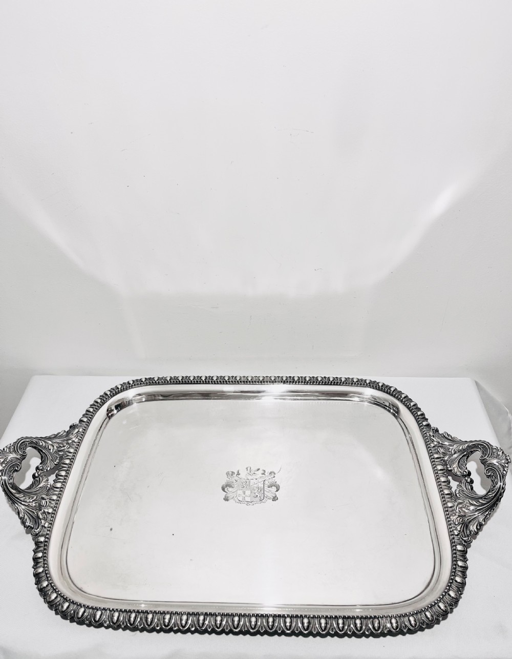 A rectangular silver-plated tray featuring ornate cast acanthus handles, a gadrooned rim, and an engraved heraldic coat of arms at the centre.