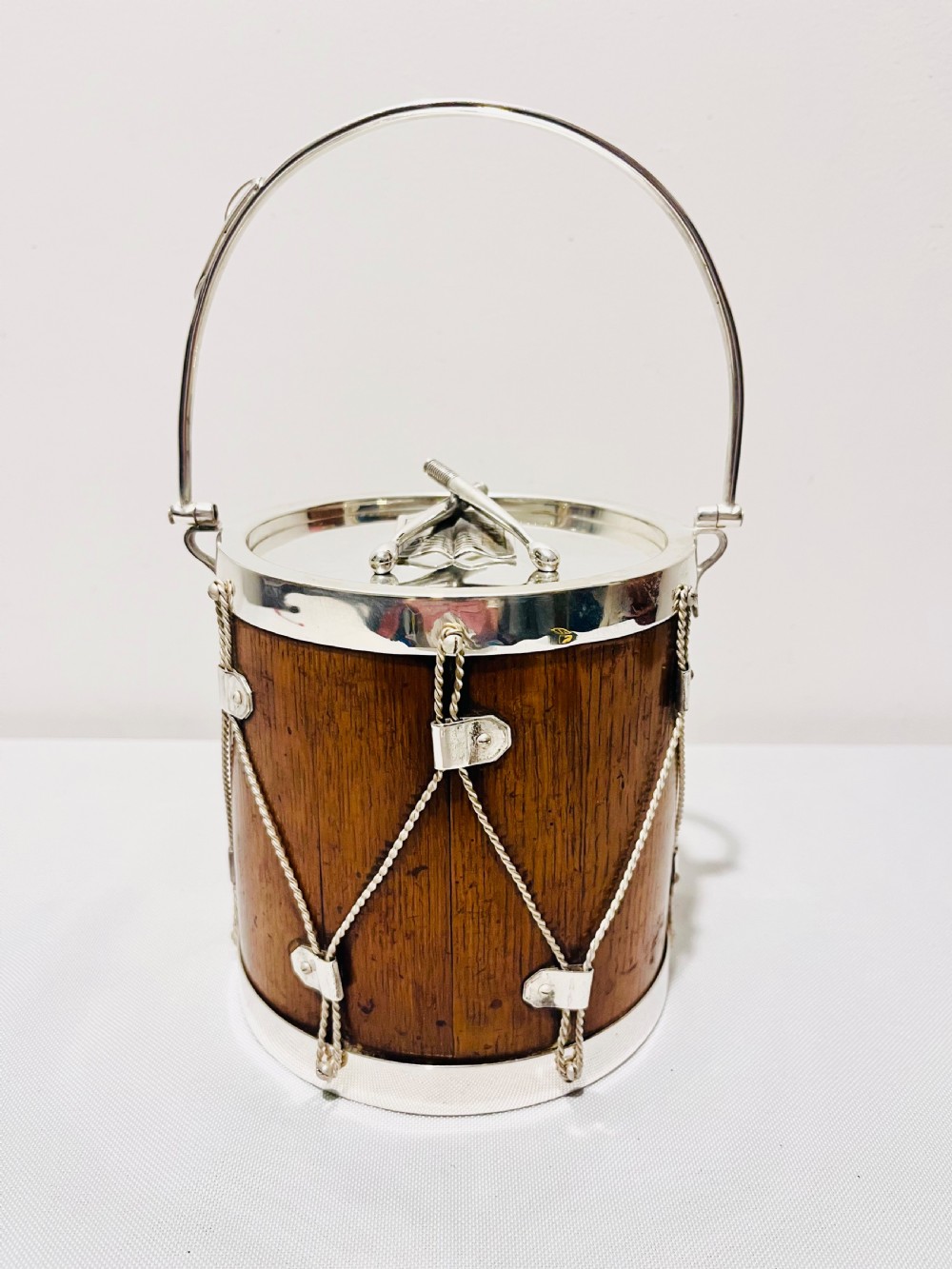 A drum-shaped antique oak biscuit box featuring silver-plated rims, rope cord detail, crossed drumstick lid finial, and a silver swing handle.