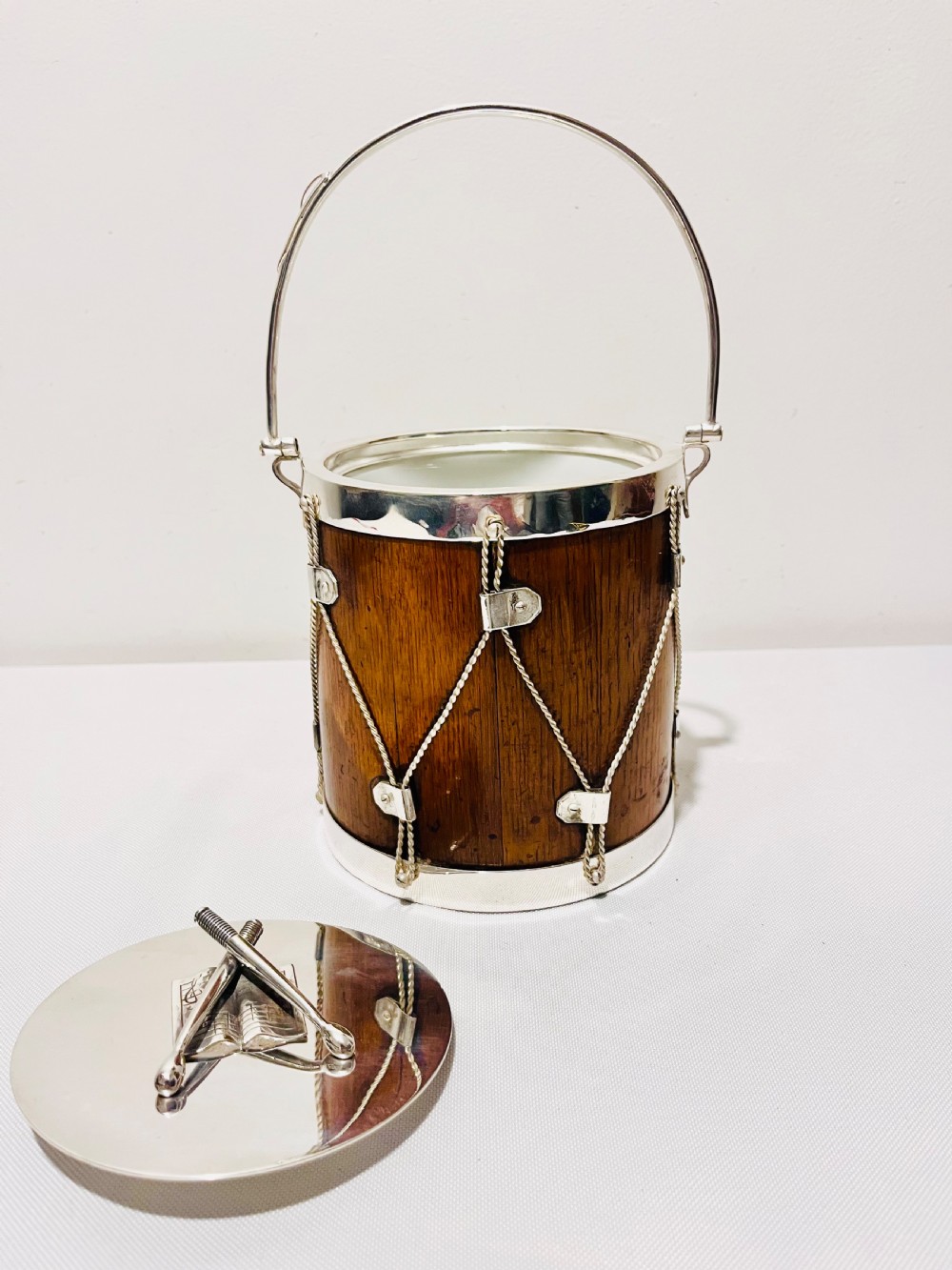 A drum-shaped antique oak biscuit box featuring silver-plated rims, rope cord detail, crossed drumstick lid finial, and a silver swing handle. View VIII