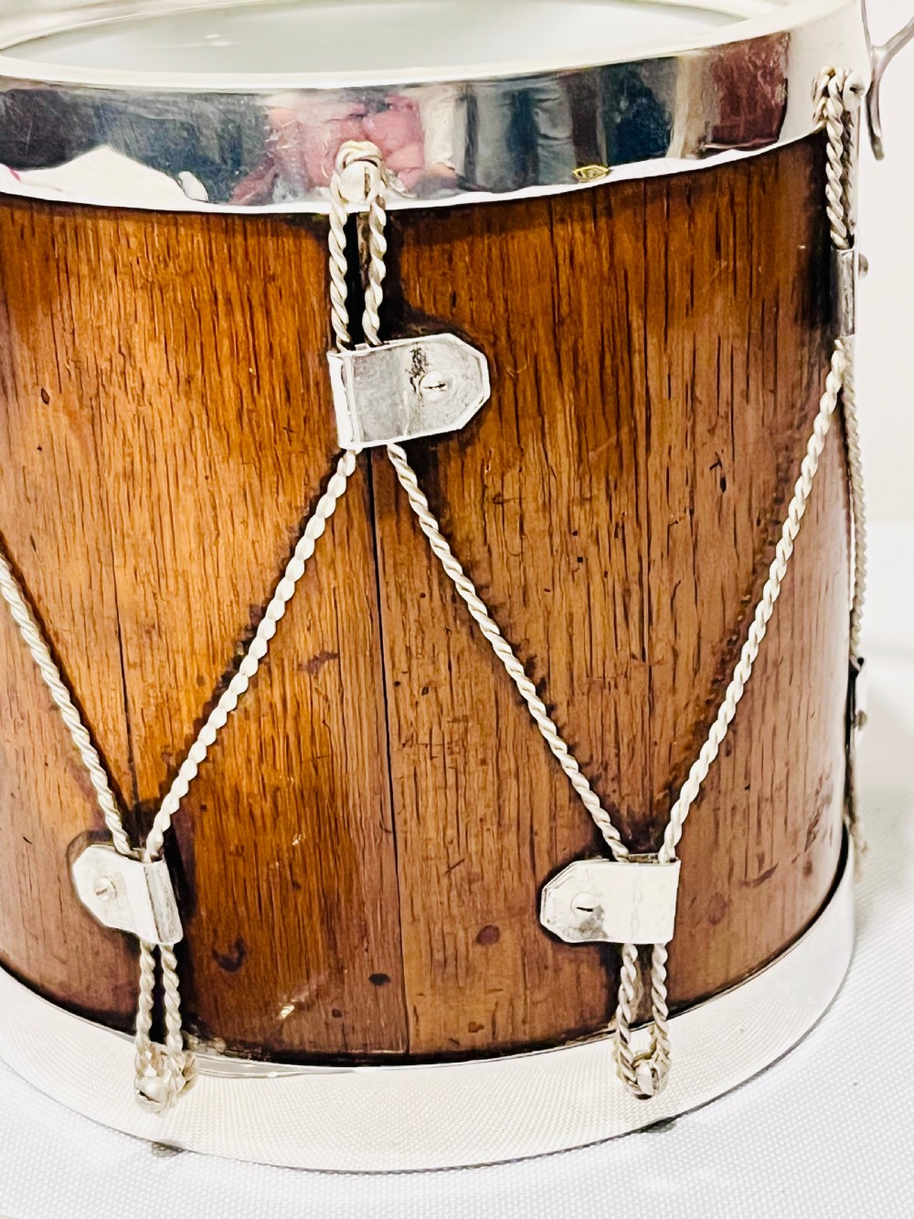 A drum-shaped antique oak biscuit box featuring silver-plated rims, rope cord detail, crossed drumstick lid finial, and a silver swing handle. View IX