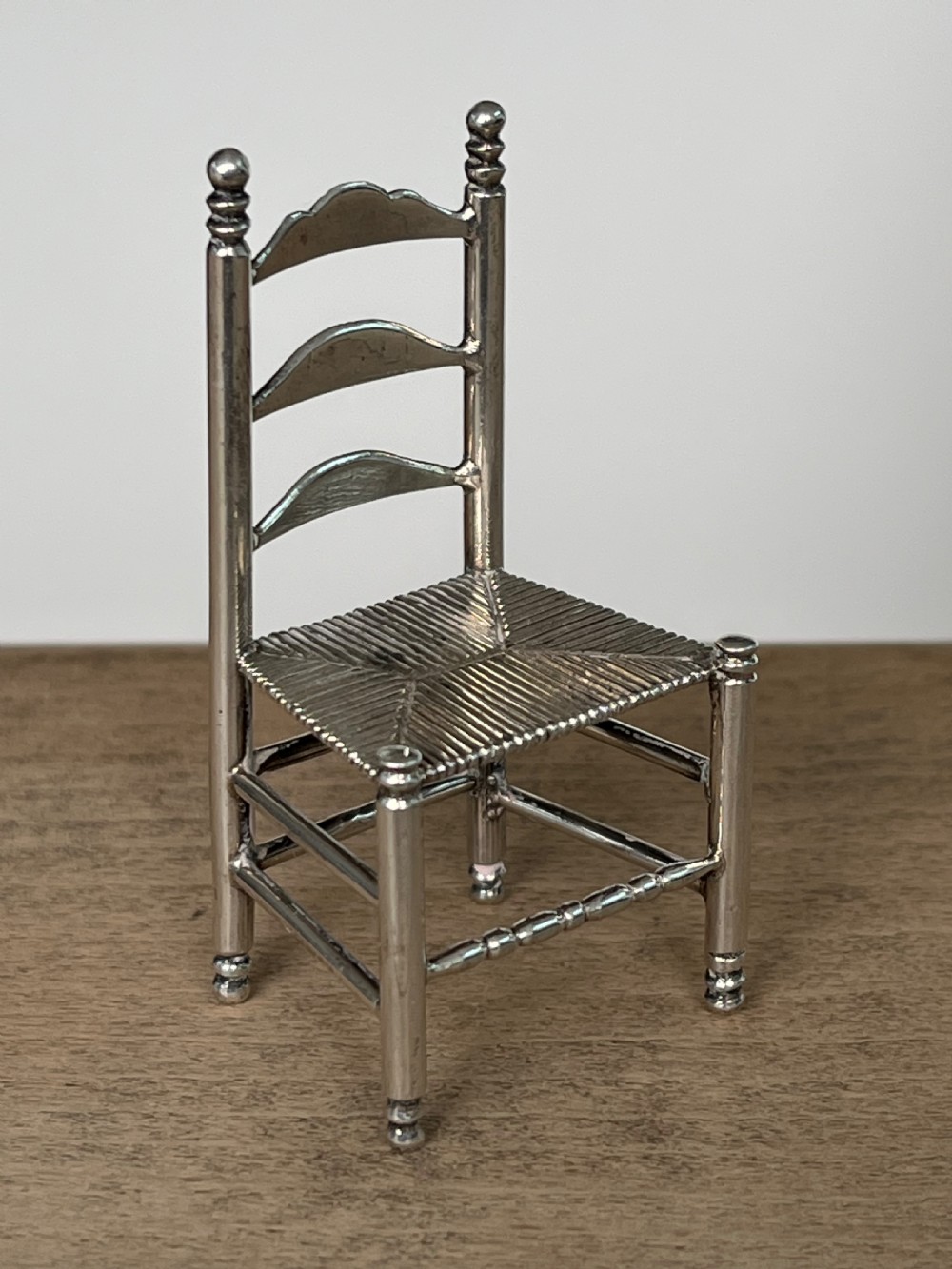 A miniature silver chair featuring a ladderback structure, turned legs topped with finials, and a textured woven seat.