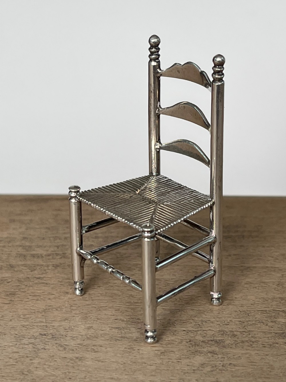 A miniature silver chair featuring a ladderback structure, turned legs topped with finials, and a textured woven seat. View III