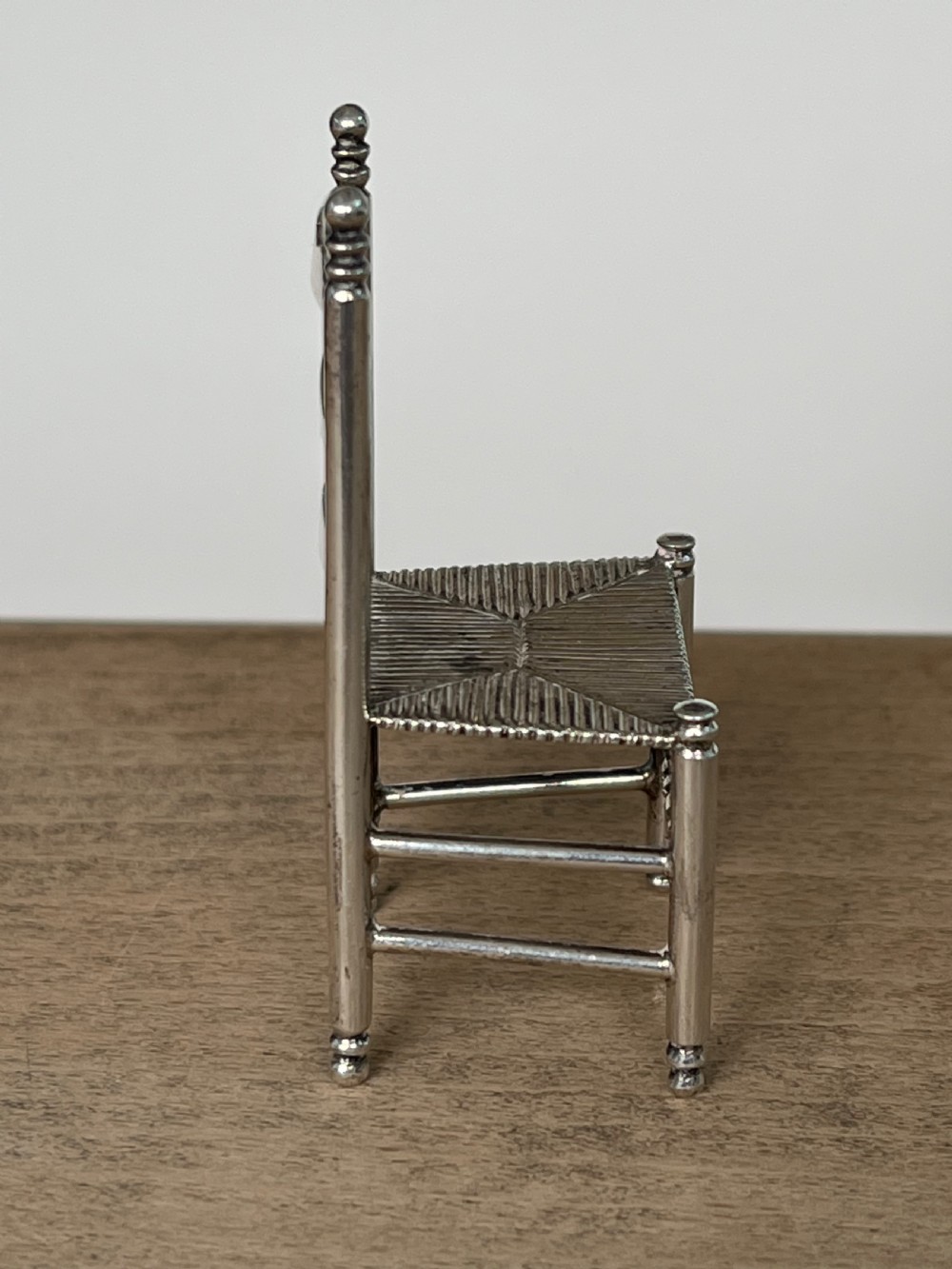 A miniature silver chair featuring a ladderback structure, turned legs topped with finials, and a textured woven seat. View IV