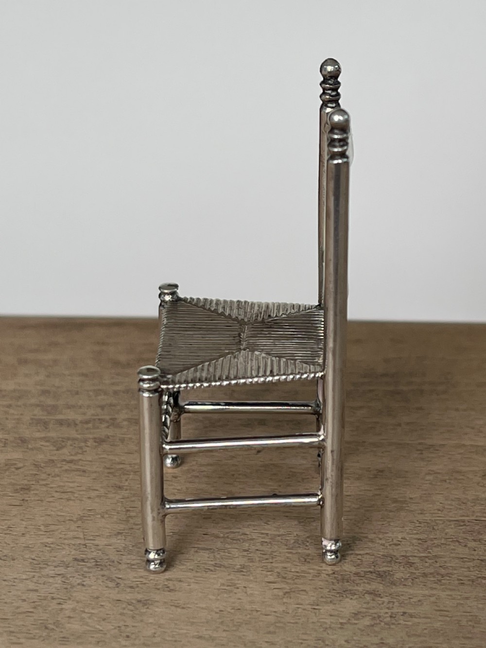 A miniature silver chair featuring a ladderback structure, turned legs topped with finials, and a textured woven seat. View V