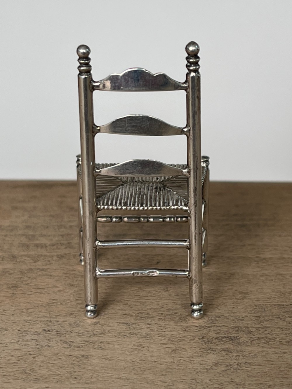 A miniature silver chair featuring a ladderback structure, turned legs topped with finials, and a textured woven seat. View VI