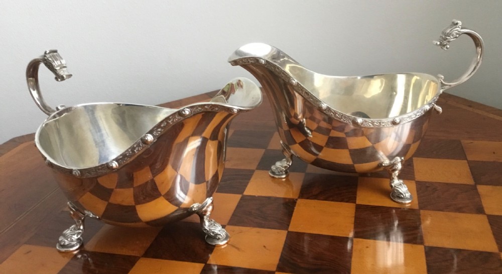 A pair of polished silver sauce boats with poured spouts, capped handles, and decorative tripod feet resting on a chequered surface.