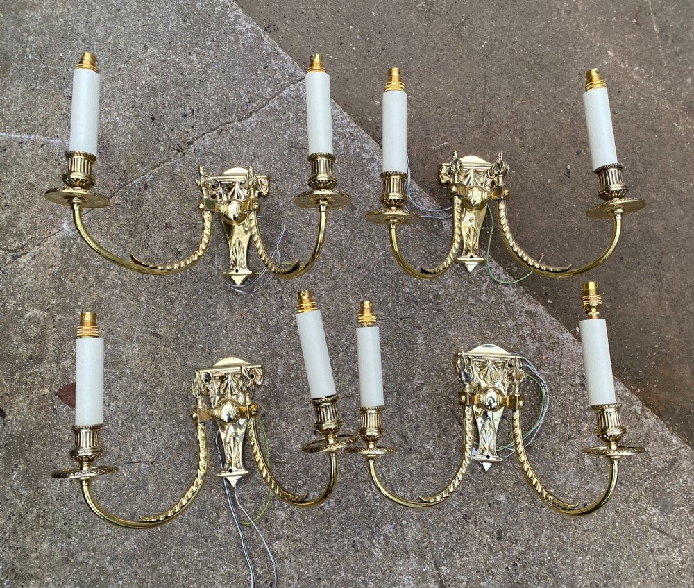 Set of four Edwardian cast brass twin-arm wall lights with reeded scroll arms, foliate backplates, and cream candle sleeves.