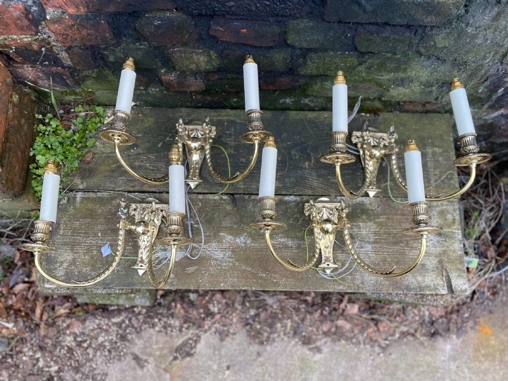 Set of four Edwardian cast brass twin-arm wall lights with reeded scroll arms, foliate backplates, and cream candle sleeves. View III