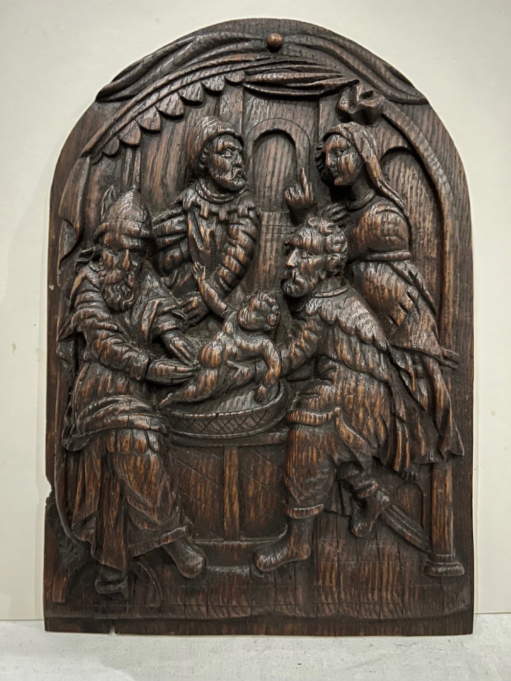 An arched dark oak panel featuring a high-relief hand carving of the circumcision of Christ with several figures.