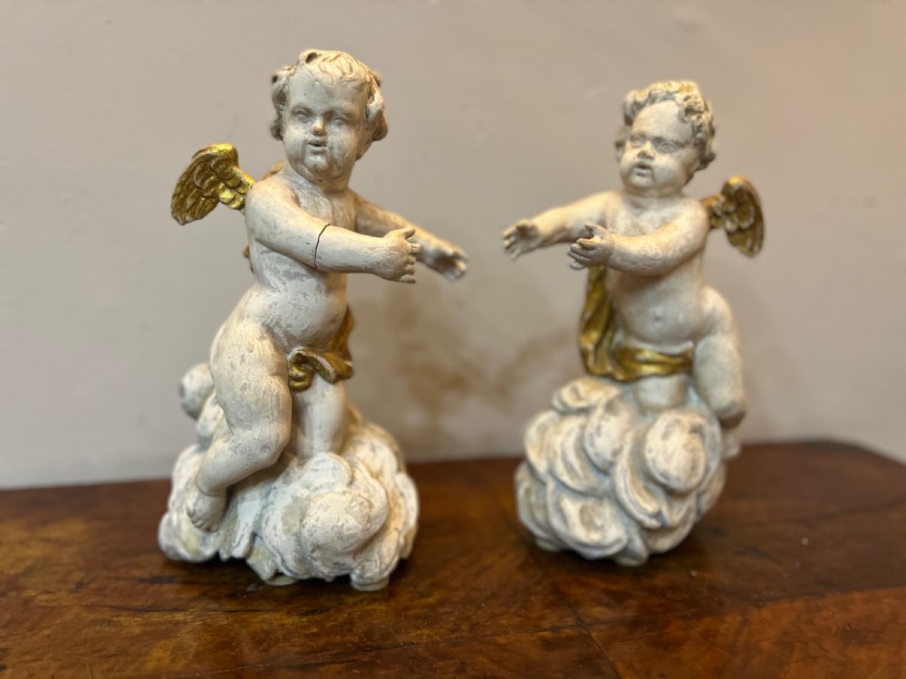 A pair of 18th-century carved wood polychrome putti featuring painted off-white figures with gilded wings and drapery, resting upon carved cloud bases.