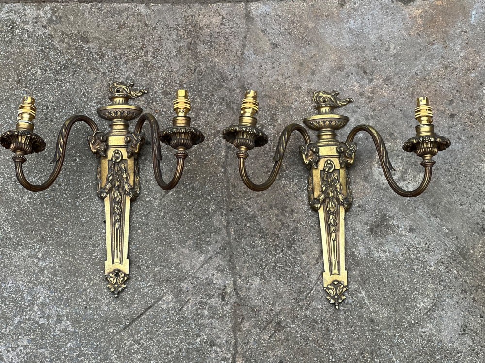 A pair of Edwardian cast brass twin-arm wall lights designed in the Neoclassical Adam style with urn finials and tapered backplates.