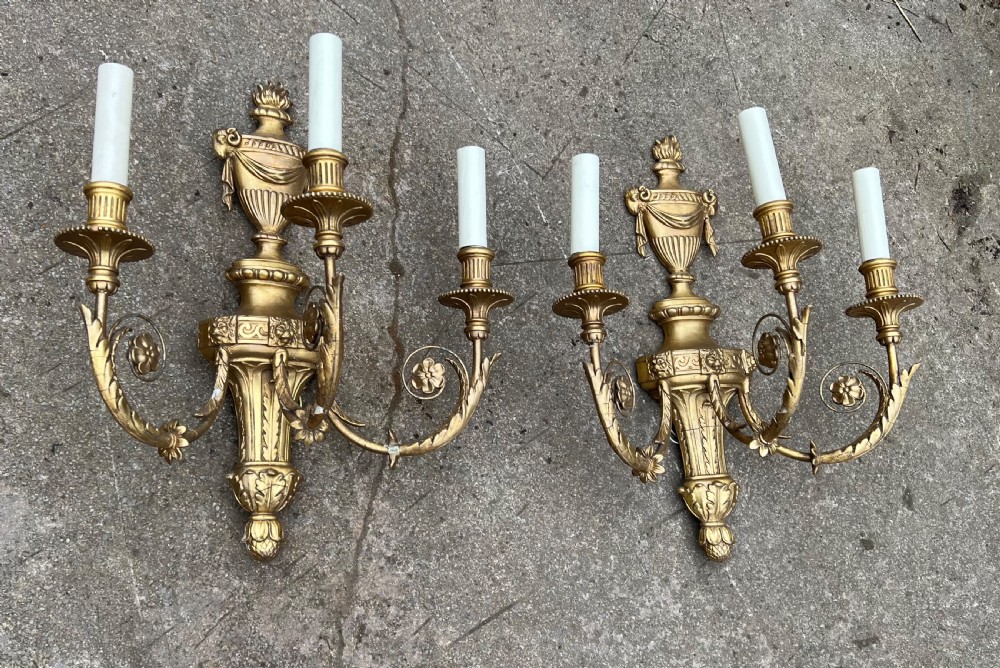 A pair of three-arm Neoclassical style giltwood and gesso wall sconces featuring urn backplates and foliage scrollwork.