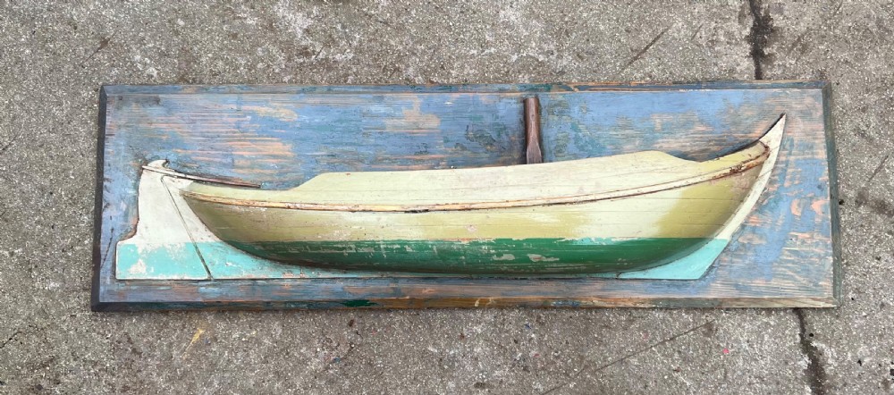 A carved wooden half-hull boat model with cream and green painted finish, mounted on a rectangular blue wooden backboard.