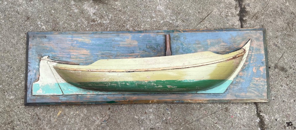 A carved wooden half-hull boat model with cream and green painted finish, mounted on a rectangular blue wooden backboard. View II