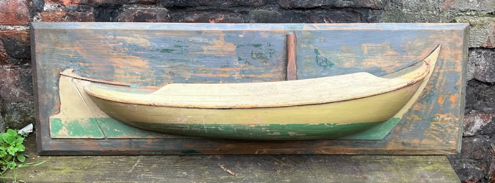 A carved wooden half-hull boat model with cream and green painted finish, mounted on a rectangular blue wooden backboard. View V