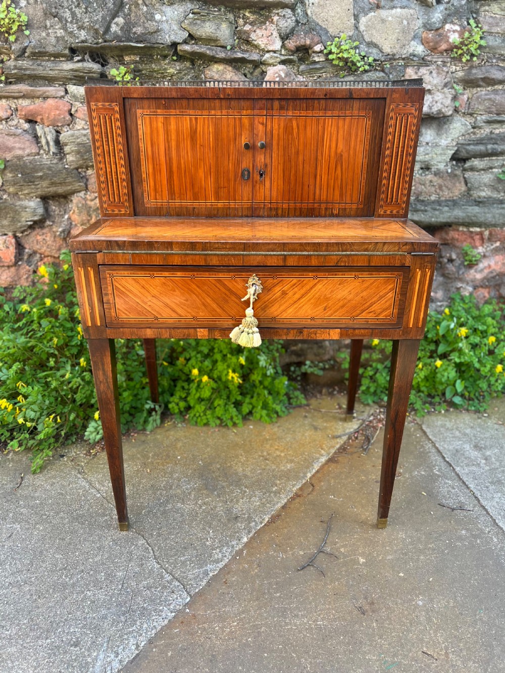 An 18th-century French bonheur de jour desk in warm brown inlaid wood, featuring square tapered legs, a single drawer with a tassel key, and a top cabinet with a pierced brass gallery.